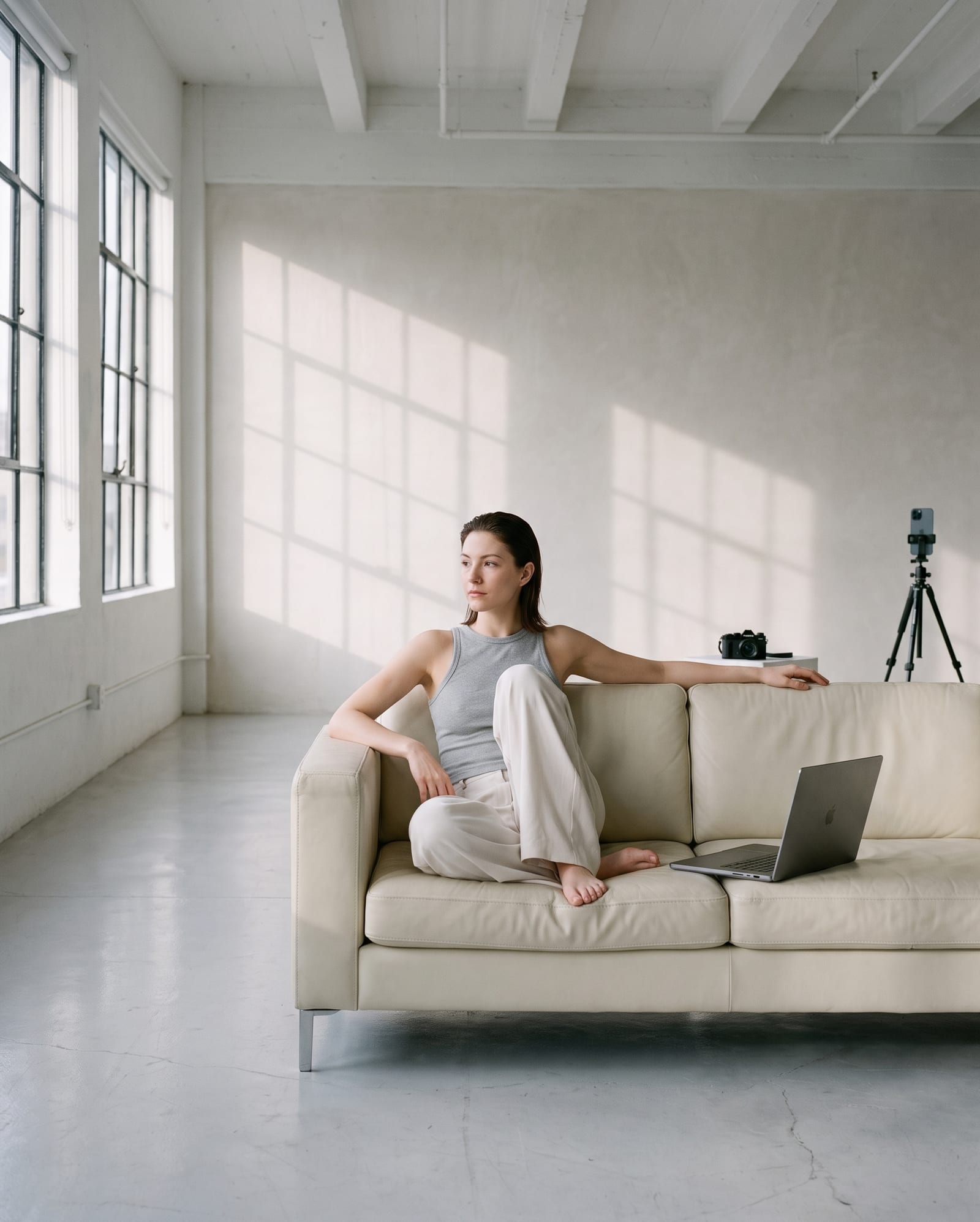 A woman on a pale sofa in a daylit loft, a laptop beside her and a phone on a tripod behind.