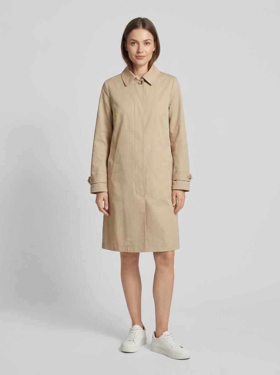 A woman in a beige trench coat standing full length on a white studio ground.